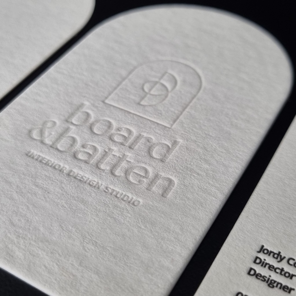 Raking light across a blind deboss pressed into uncoated white card, the paper grain visible in the shadow.